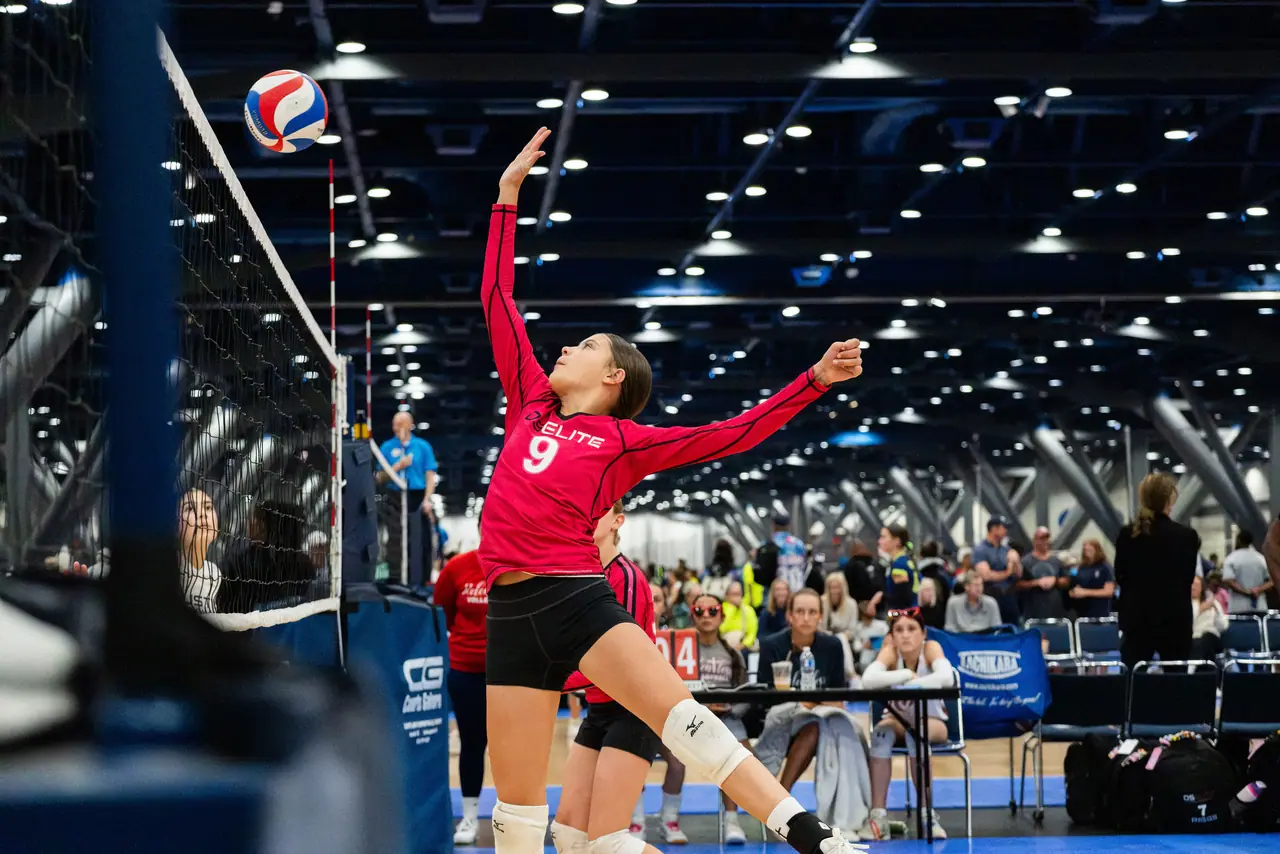 DS Elite player in a raspberry long-sleeve uniform tossing the ball to serve under the lights of a tournament hall